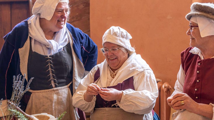 Three people in Tudor costumes are laughing together against a peach wall. There is a bunch of lavender in the foreground.